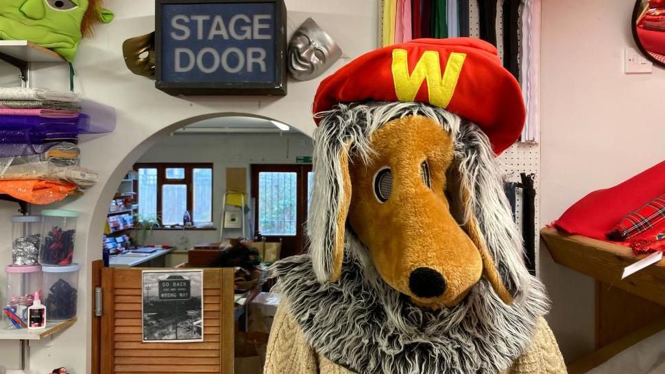 The head of a Womble costume. A cream jumper is peeping out from below the head. A sign in the background says "stage door".