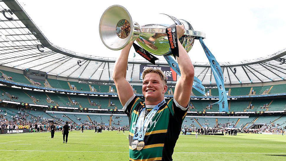 Northampton flanker Henry Pollock, wearing the club's yellow, green and black shirt, lifts the Prem Rugby trophy above his head