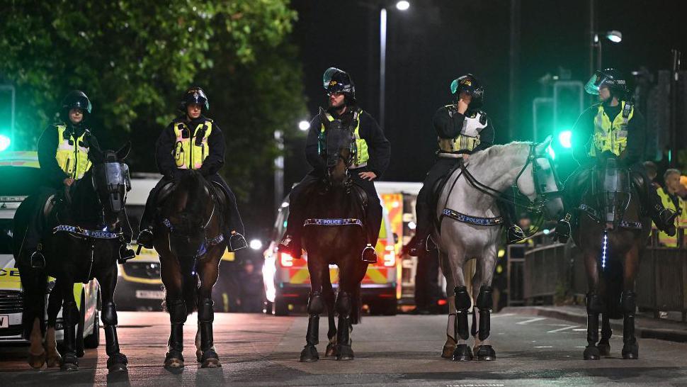 Five police officers on horses and wearing riot helmets stand in line across street at night