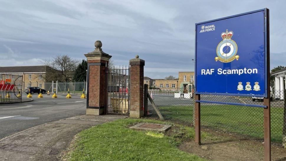 A blue RAF Scampton sign outside the former air base. Some of the brick buildings can be seen behind the fence. There is grass and a white car parked in the distance.