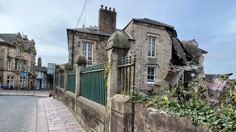 A three-storey grey-stone building has partially collapsed. Half of the building is standing while the other half is largely missing with pipework and stone poking out of it, In the foreground, there is a green metal railing.