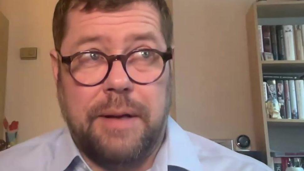 Graeme Stewart in a screengrab from a Zoom interview. He sits in a room with some bookcases behind him, and wears a smart shirt and glasses.