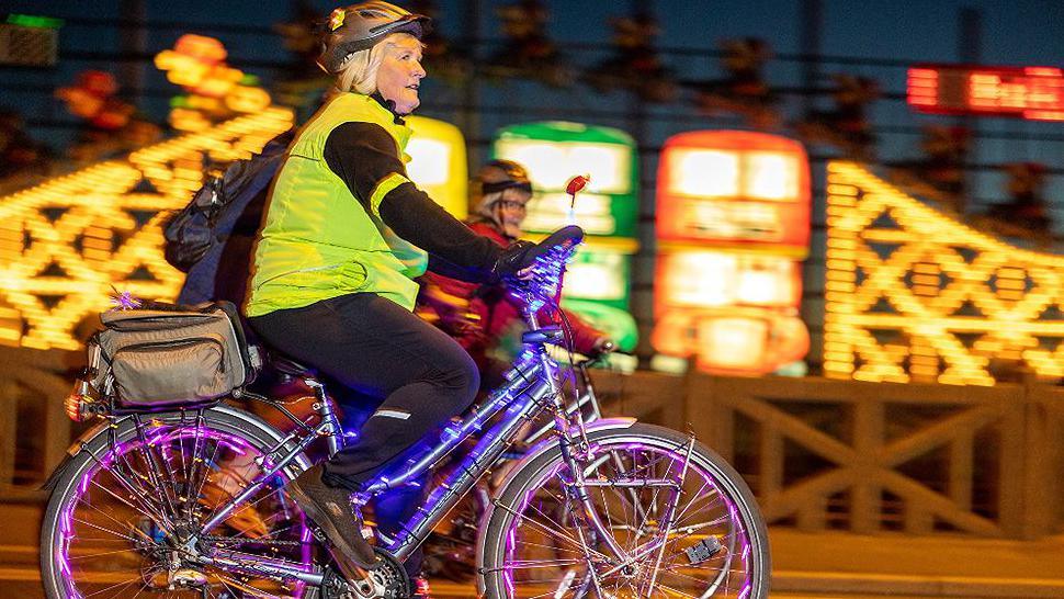 A woman cycling past bright lights wearing a helmet and high vis jacket on a bicycle with purple lights on the inside of the wheels