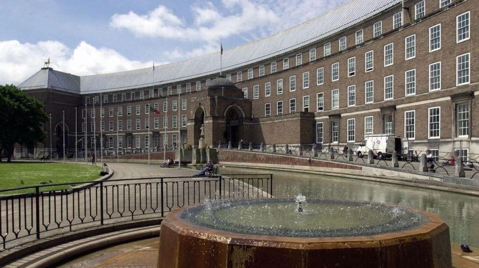 The Bristol City Council building which is a curved, brick municipal building with a fountain outside and railings in front of a green space.