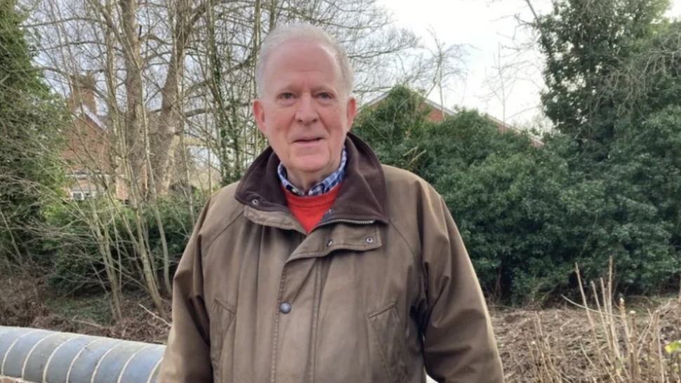 A man with short white hair stands in front of trees and bushes. He is wearing a blue check shirt, a red pullover and a brown coat.