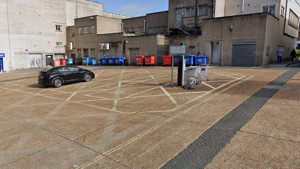 The Marks & Spencer Rear Car Park has one black car parked in it. It is lined with industrial bins and the backs of shops. There is a row of shopping trolleys parked next to the car park sign.