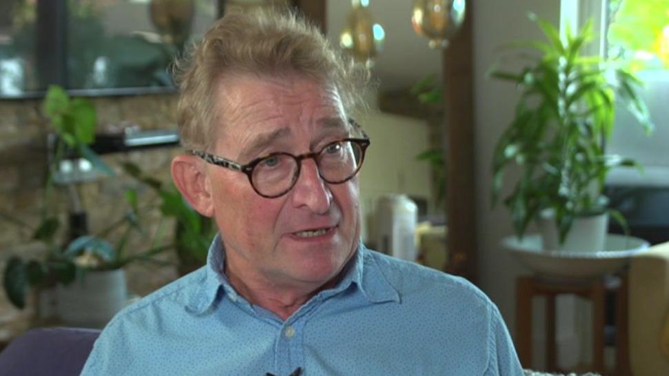 Dr Chris Clough being interviewed. He is wearing glasses with dark rims and a blue shirt. There are plants behind him, sat on small tables, with a TV on a wall.