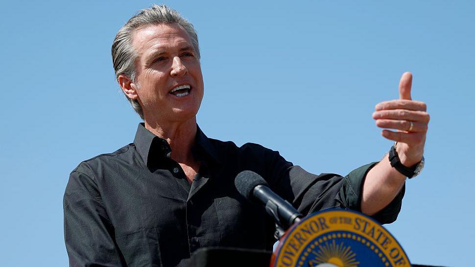 Gavin Newsom speaks at a podium outside with blue skies behind him. He is wearing a black shirt and gesticulating with his left arm