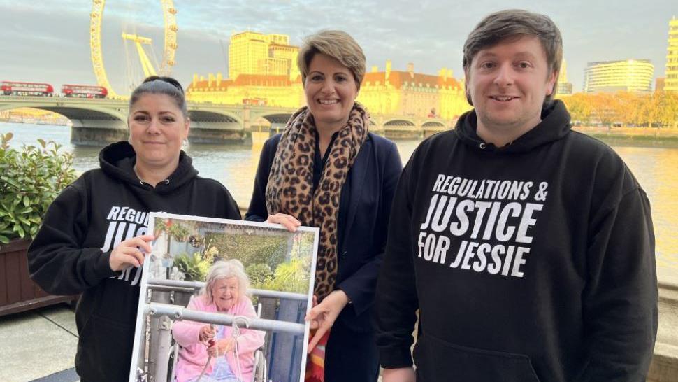 Claire Stockdale, Emma Hardy MP and Tristan Essex smiling and holding a picture of Jessie Stockdale. Tristan and Claire are wearing black hoodies with 'regulations and justice for Jessie' written on the front. You can see London red buses and the London Eye in the background.