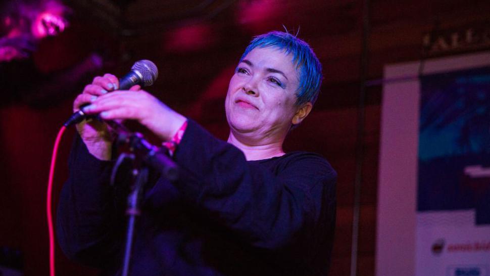 Leslie Rankine has short hair which is either dyed blue or lit up blue by stage lights. The rest of the image is lit in red. She is about to sing into a microphone