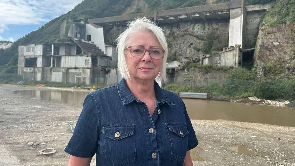 Tina Brennen, who had blonde hair in a short bob and is wearing glasses and a blue denim short-sleeve shirt, standing on the Summerland site. The derelict remains of the Aquadrome are in the background behind a large puddle in the gravel.