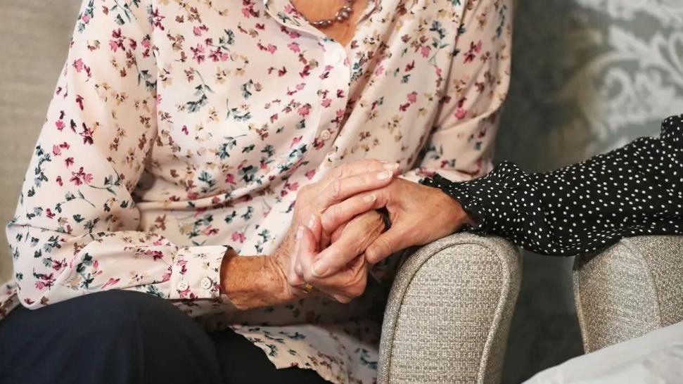 A close-up of the hands of two women sitting on armchairs holding hands. One has a black blouse with white spots, the other wears a pink blouse with a flower design.
