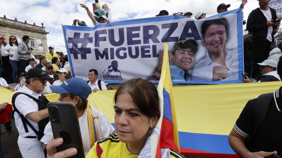 People demonstrate with signs and flags during the "March of Silence" in Bogota, Colombia, 15 June 2025. A woman, wearing the shirt of Colombia's national football team, can be seen holding up a mobile phone. Behind her, people are carrying a banner reading "Strength, Miguel".