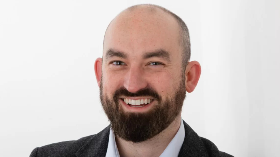 A headshot of Eoin Hayes. He is a balding man with a dark beard. He is posed up against a white background. He is wearing a white collared shirt and a dark suit jacket.