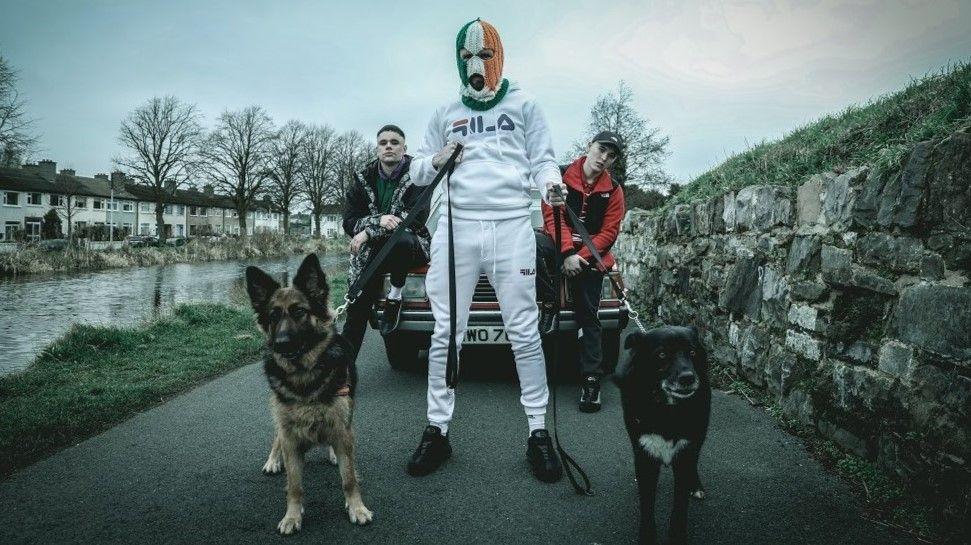 A member of Kneecap in a white tracksuit and a green-white-and-orange balaclava standing with two dogs in a lead in front of fellow band members sitting on a car