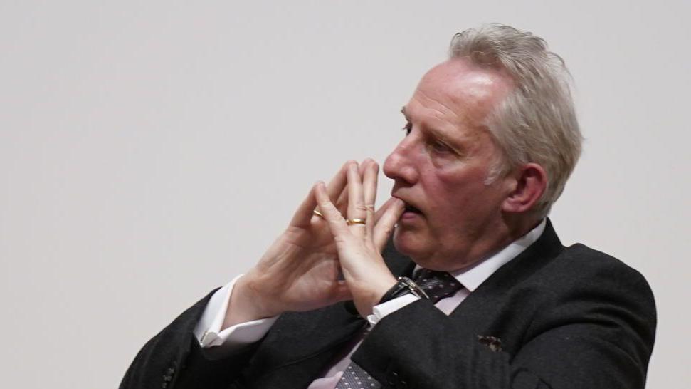 A man in a dark suit, with grey hair is sitting with his hands together up to his face in a gesture of consideration. The photograph is taken from a side angle. 