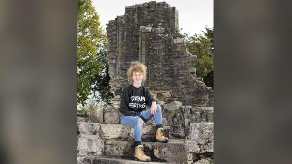 Rebekah Watkins, in jeans, brown walking boots, and black sweatshirt reading Dream Heritage, is sat on the stairs of the manor house. She is smiling at the camera. Her blonde, curly hair is put in a bun. 