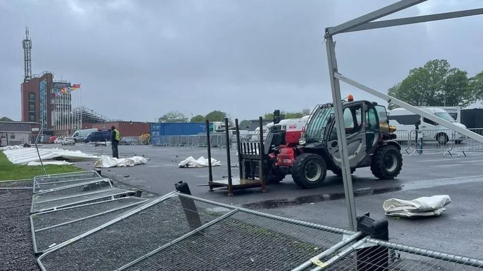 Wet tarmac in the TT paddock area with temporary fencing and tents blown over, along with a parked red, grey and black HGV and several parked vans. A man dressed in black wearing a yellow hi-vis vest stands looking a the damage with the TT grandstand and tower in the background against the backdrop of a grey cloudy sky.