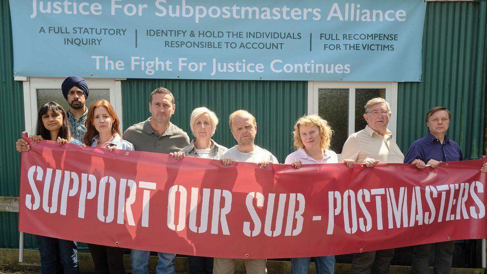 ITV drama Mr Bates vs the Post Office scene showing cast members portraying real life postmasters - with Toby Jones centre as Alan Bates, and Ifan Huw Dafydd second from right as Noel Thomas from Anglesey