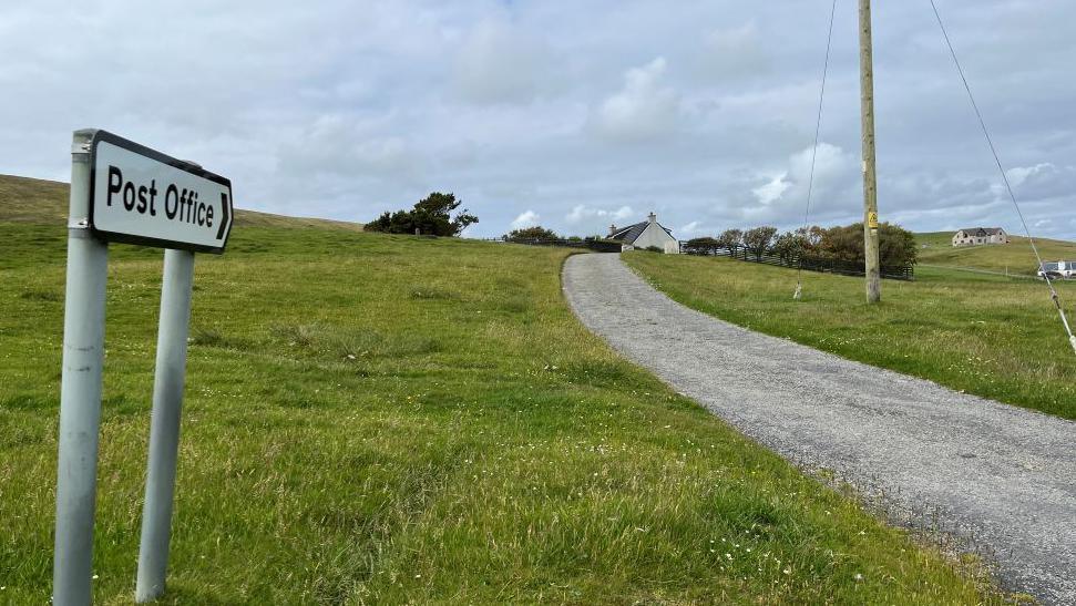 A road sign pointing to the Post Office. It is mounted above a grass verge, next to a narrow road. A house and trees are visible on the horizon.