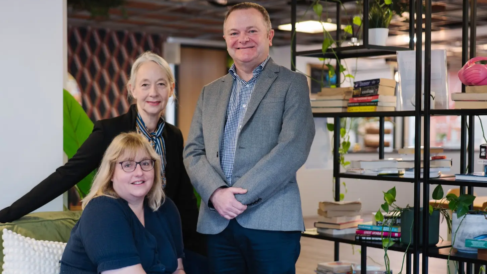 Three leaders of the BID, Vivienne, Anna and Steve are in a light office with an open-shelved bookshelf behind. Vivienne is seated with the others standing behind her. They are all smiling to camera and wearing smart business clothes.