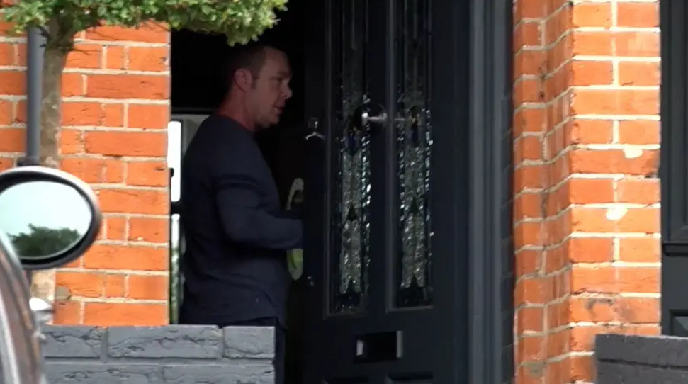 A photo of a man in the doorway of a red brick house. He is standing side on and wearing a blue jumper