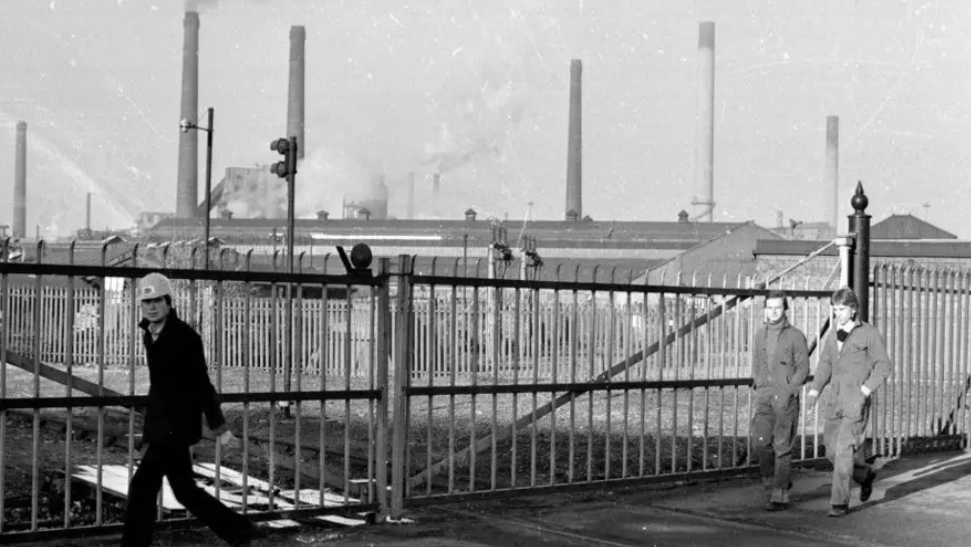 A black and white photo of Corby's steelworks. It is a huge factory with several chimneys. It is in the distance behind a fence. A man in a hard hat looks at the camera as he walks in front of the fence. Two other men in boiler suits are walking past a short distance behind him.