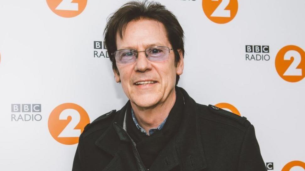 Shakin' Stevens pictured in 2021. He has short dark hair and is wearing glasses. He is smiling at the camera and is wearing a black jacket. He is pictured from the chest up, in front of a BBC Radio 2 billboard.