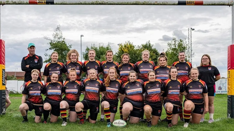 Two rows of rugby players stood under the posts with coaches either end of the rows. Their kit is black, red and yellow. A white rugby ball is on the grass in front of them