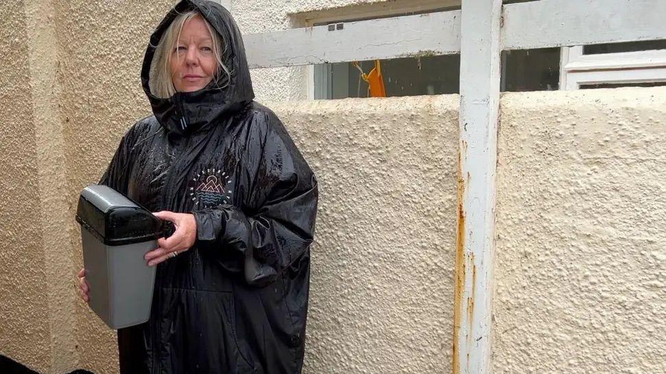 Becci Hey is pictured. She is wearing a black raincoat with the hood up and she has blonde hair. She is holding a small sanitary bin and is standing outside a cream-coloured toilet block.