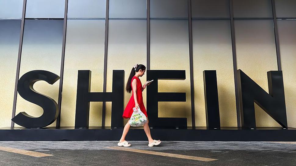 A woman in a red dress looks at her smartphone while walking past a Shein sign outside the company's office in Guangzhou, China. 