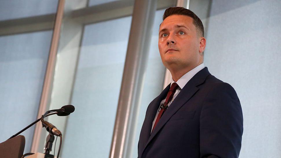 Wes Streeting stood at a lectern delivering a speech. He is wearing a navy blue suit, pale blue shirt, and red tie. 