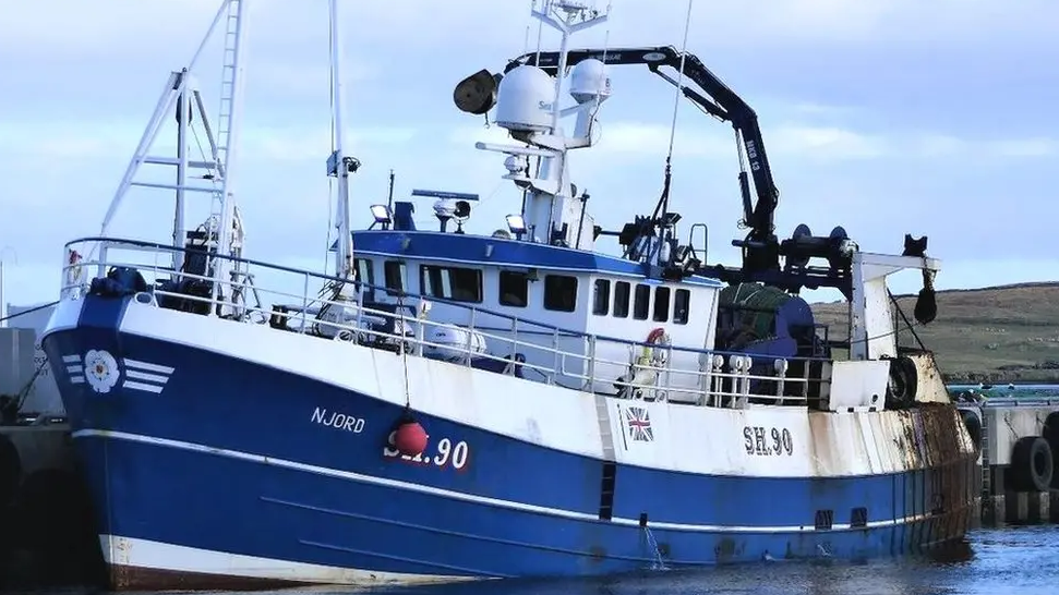 The Njord boat. It is mainly blue on the hull with a white deck. It is sitting on the water in front of a grey sky.