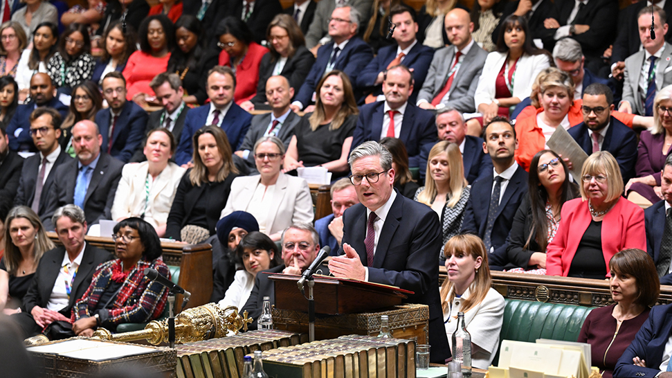 A wide shot of the Labour benches in the Commons chamber, with Sir Keir Starmer speaking at the despatch box and the Parliament mace and Erskine May books in the foreground