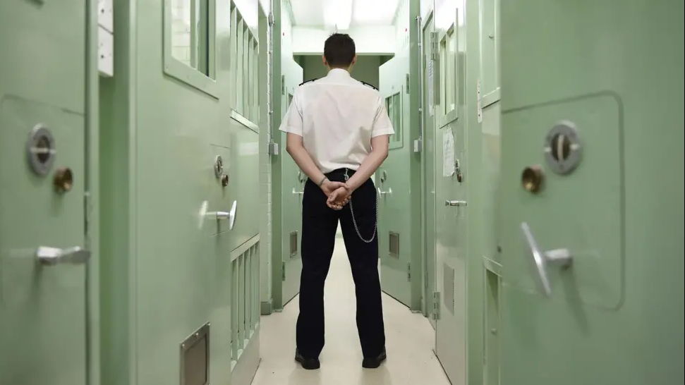 A male prison officer standing in a prison, in uniform, with his hands behind his back and his back to the camera. He's standing in a narrow corridor between two rows of cells with light green doors.