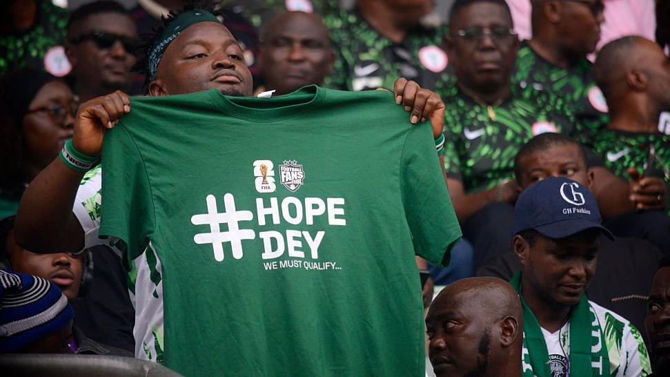A Nigeria football fan holds a green t-shirt with the words "Hope Dey" (meaning there is hope) written in white, and "we must qualify" underneath. He is surrounded by other Nigeria fans who are seated in a stadium 