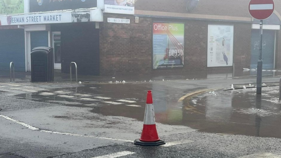 Flooding on a street in Grimsby. A lone traffic cone stands at the edge of a large pool of water. Freeman Street Market hall is next to the pool of water.