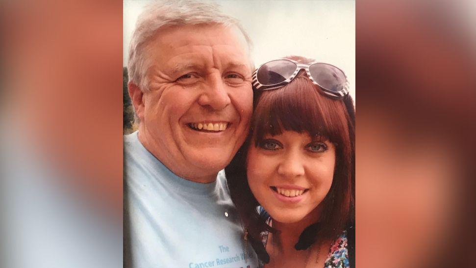 Rhian with her dad Wayne, she has shoulder length red hair and a fringe, she is wearing zebra print sunglasses on her head. They are both grinning. Wayne has short grey hair and is wearing a blue t-shirt. 