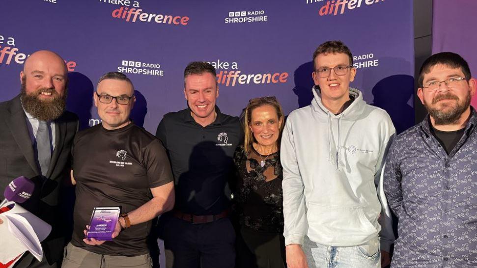 Five men and one woman standing in front of a purple background, with one of the men holding a small purple award