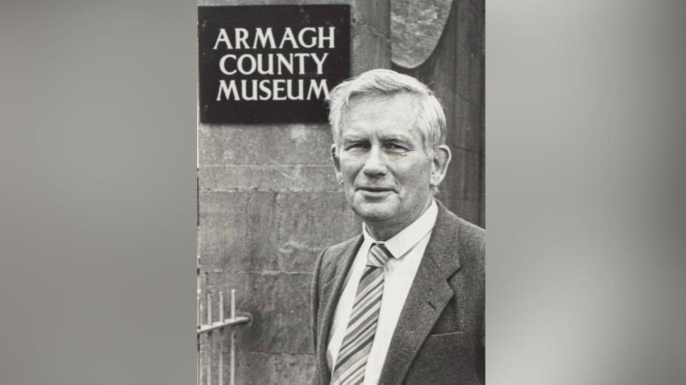 A black and white photograph of Roger, who is dressed in a suit and wearing a stripy tie. He is standing in front of a sign that says Armagh County Museum.