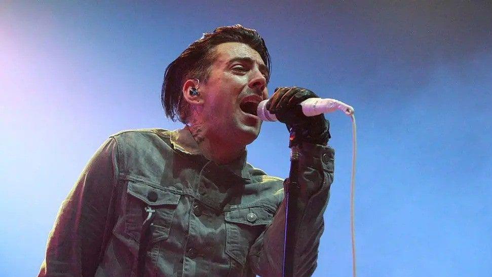 Ian Watkins, performing an outdoor gig. He is wearing a denim jacket and black gloves and is holding a white microphone.
