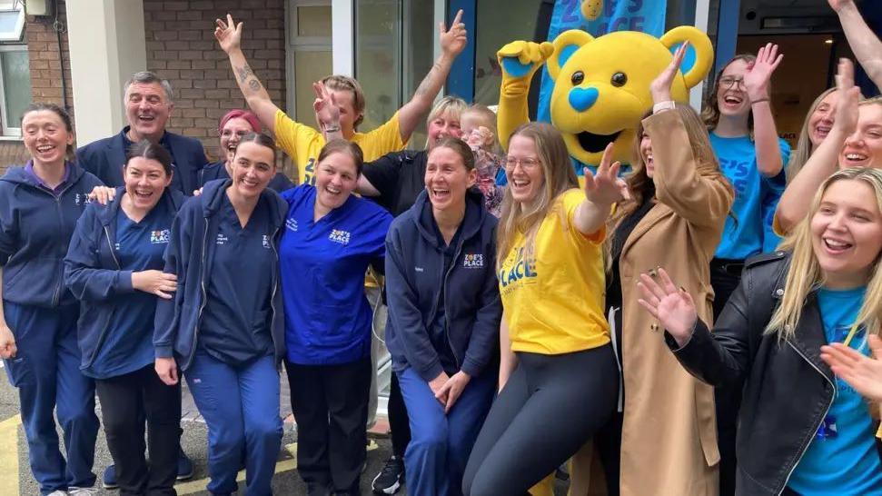 A group of nurses, some in uniform, and fundraising staff in blue and yellow Zoe's Place branded T-shirts as well as someone in a yellow teddy bear costume. They are all smiling and some are waving their arms.