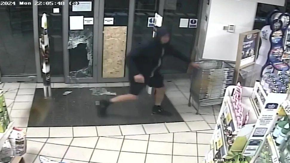 Guy Sullivan wearing black shorts and a black hoodie inside Tesco Express running away from a smashed door, with glass on the floor