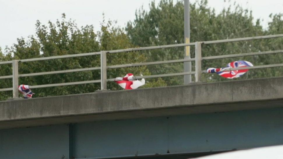 A bridge over a road with several St George's and union jack flags attached to the railing. Green trees can be seen in the background. The sky is grey.