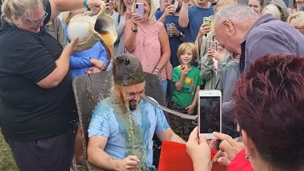 Aaron Sanderson during the coronation. He is wearing the rusty and damaged crown and a woman is pouring a large jug of beer over his head. He is surrounded by people watching on, smiling and filming the ceremony on their phone.