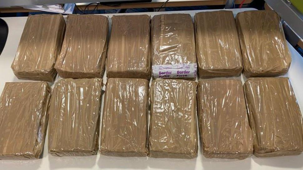 Bricks of cocaine wrapped in brown tape laid out in a row on a white table. A Border Force sticker can be seen on one of the wraps. 