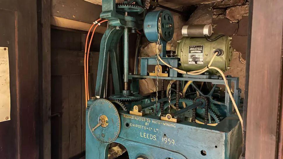 A clock mechanism with visible gears, chains and an electric motor. It’s painted blue-green.