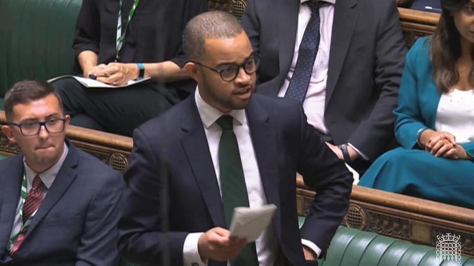 Adam Jogee, wearing a blue suit jacket, a green tie and white shirt, stands and speaks in the House of Commons while holding a piece of paper in his right hand.