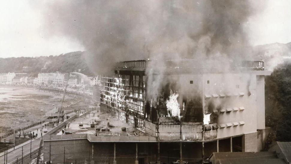 A black and white image of the Summerland complex on fire with Douglas Bay on the left.