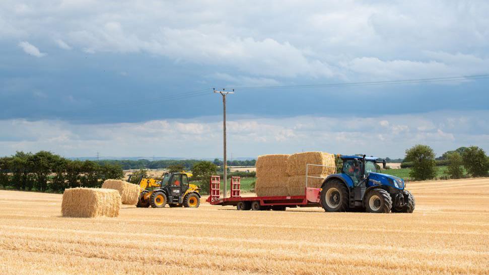 A tractor pulling a trailer with eight large hay bales on, and a second tractor helping move the hay around, in a large hay field below blue sky.